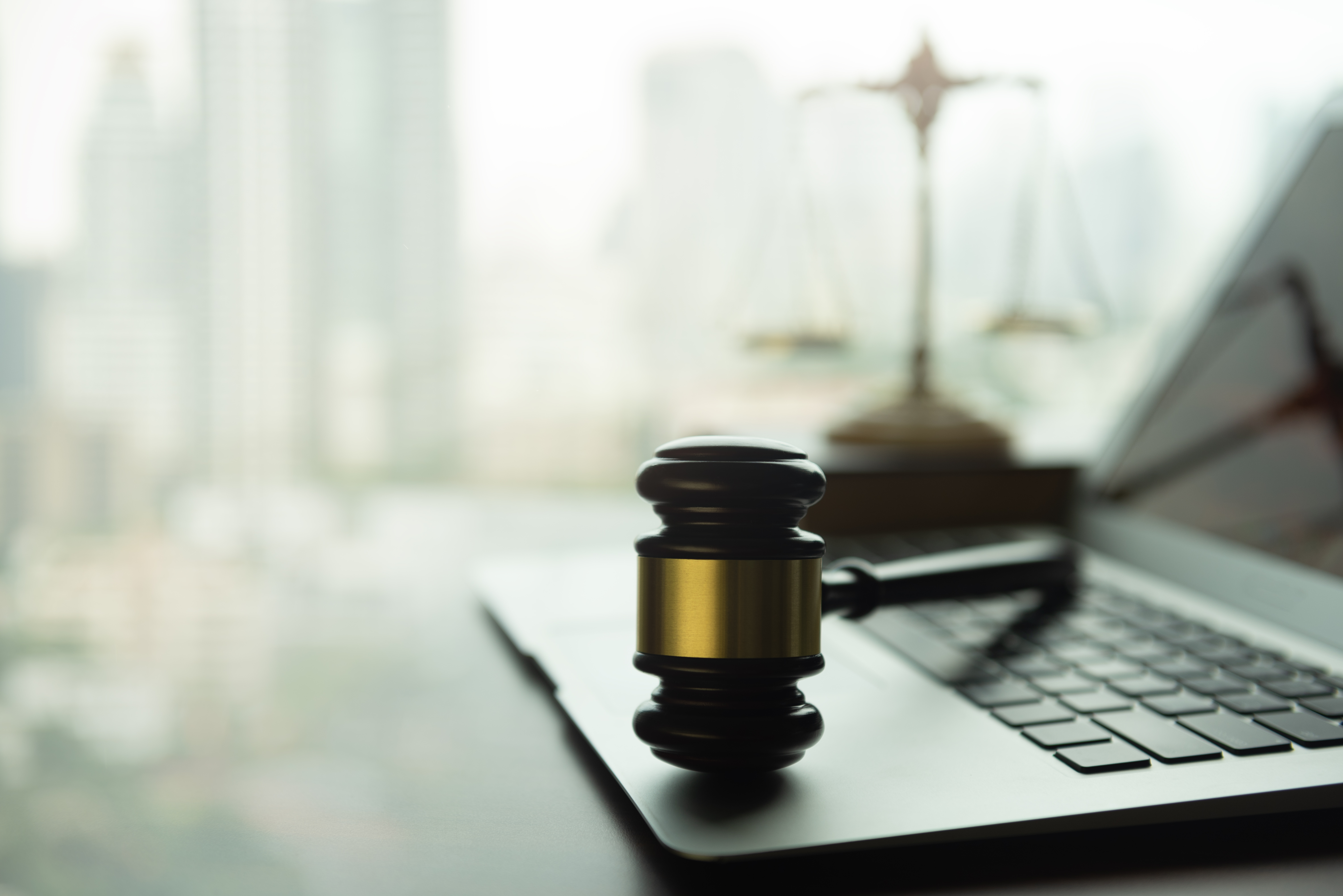 A gavel resting on a laptop keyboard with a blurred background showing a balance scale and a cityscape.