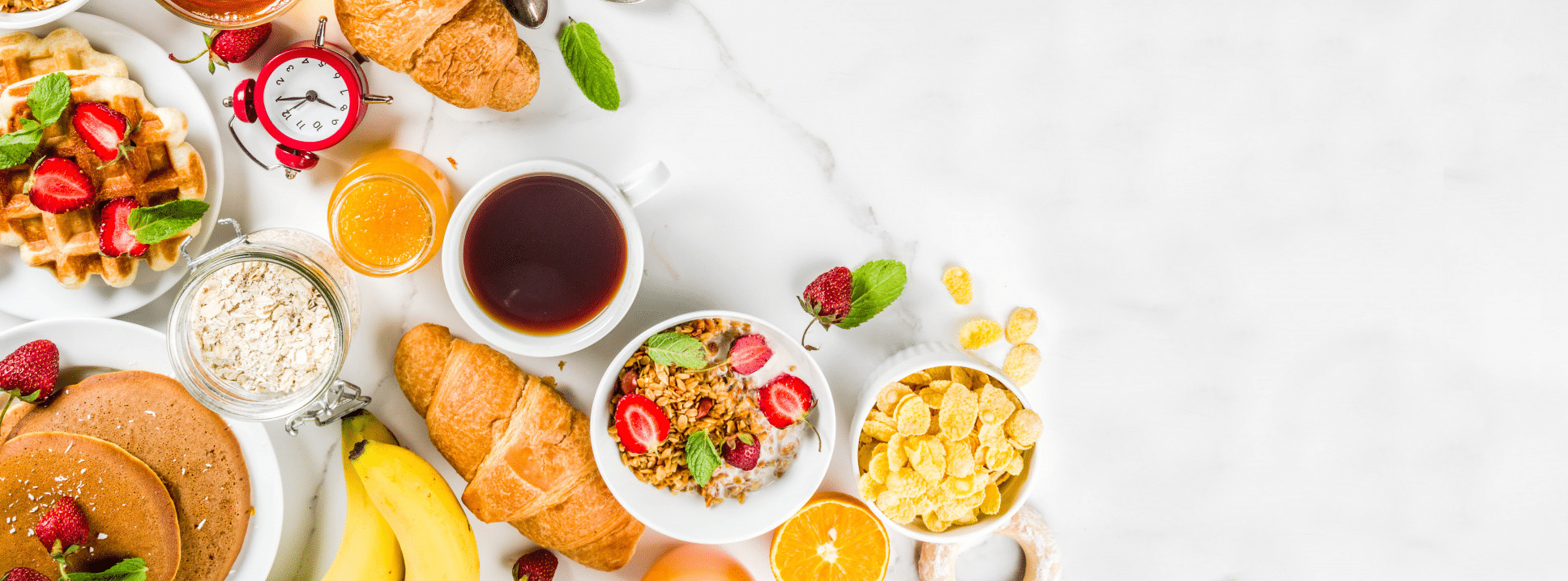 Assortment of breakfast items including waffles with strawberries, croissants, pancakes, cereal, fruit, and a cup of coffee, arranged on a white marble surface.