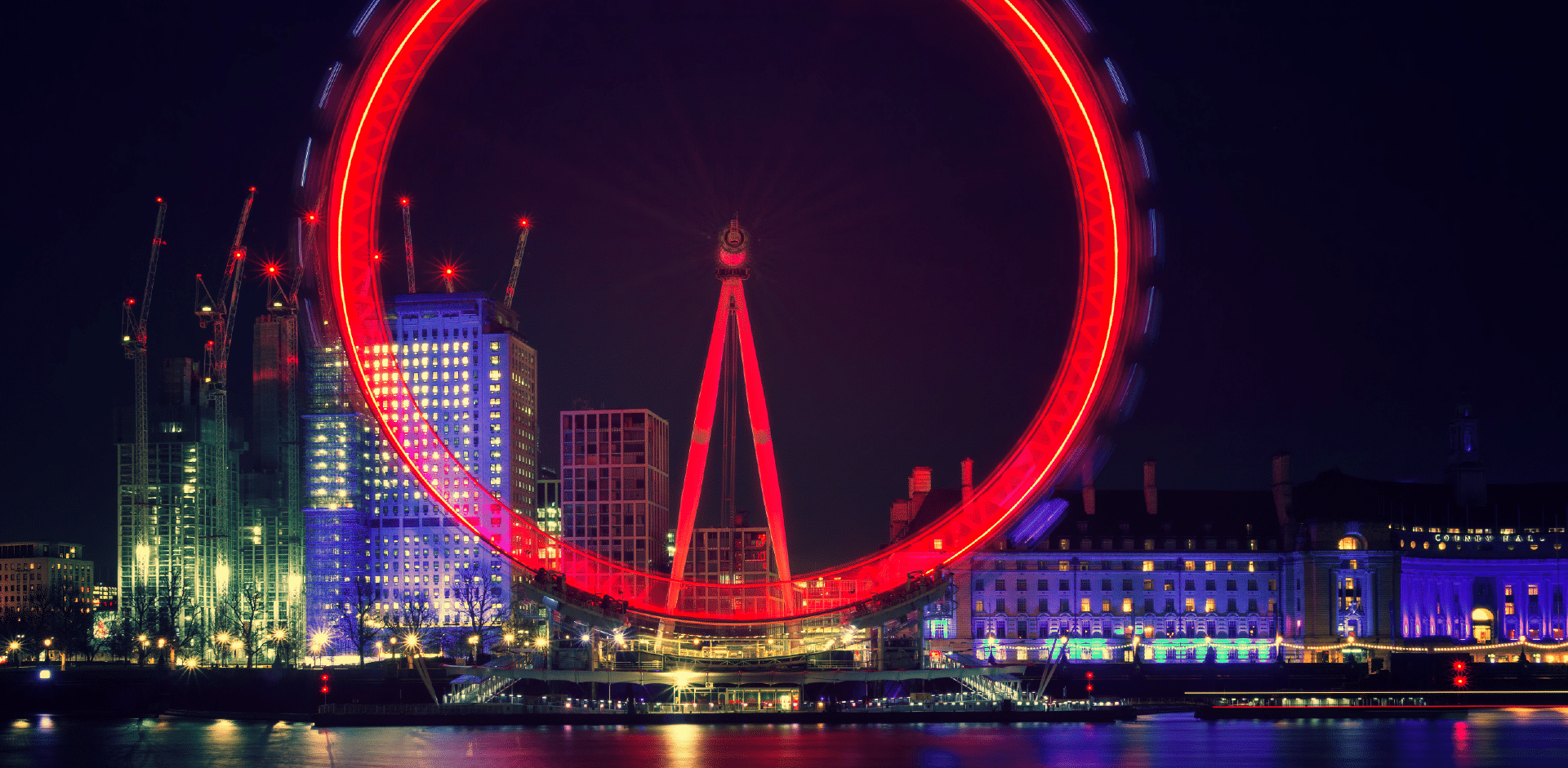 Nighttime view of the London Eye illuminated in red, with the city lights and reflections on the River Thames.