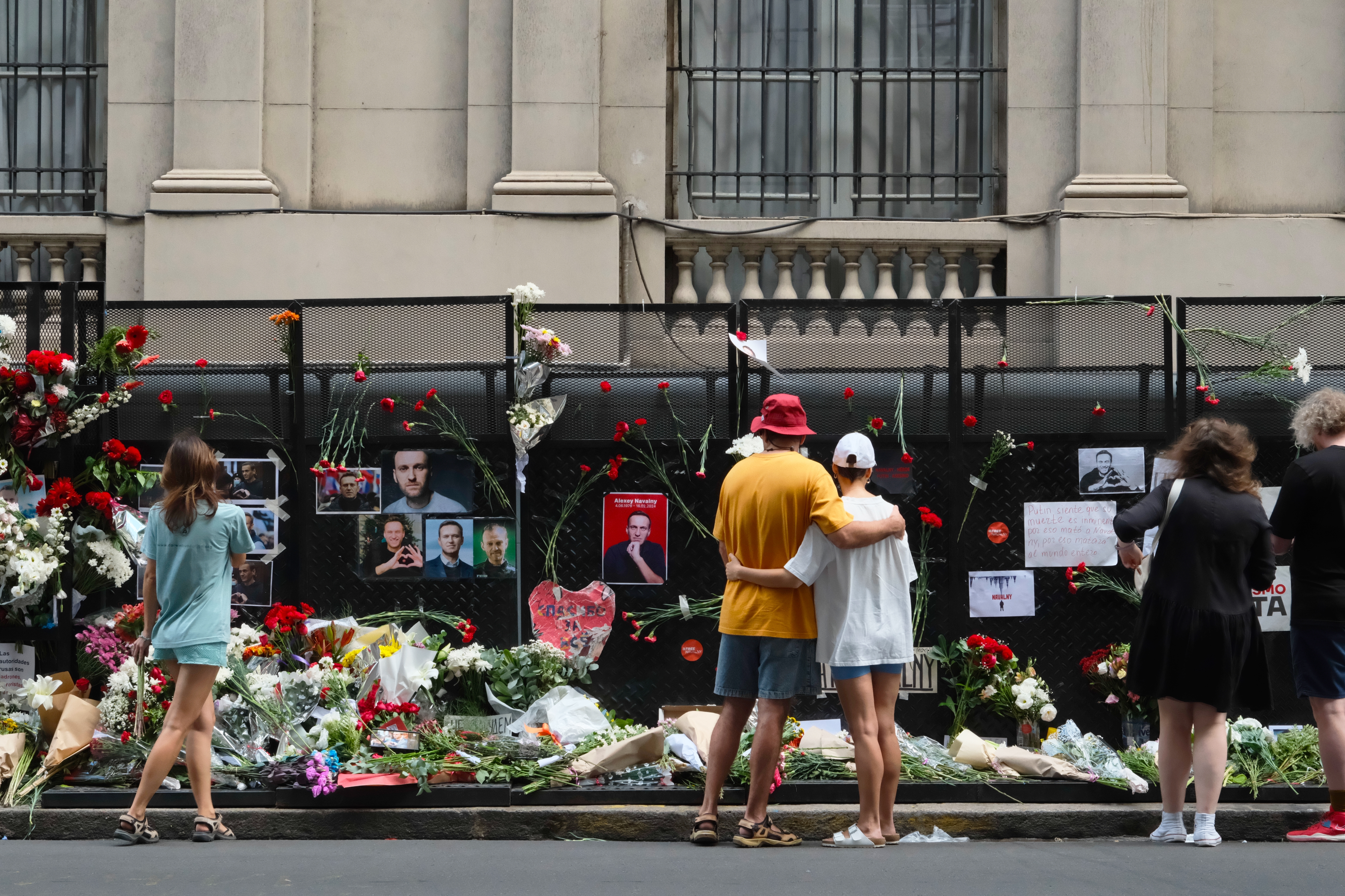 People gather and place flowers and memorials in front of a fence adorned with photos and tributes for Alexei Navalny, following the announcement of his death on February 16, 2024. The scene reflects a somber and respectful atmosphere as individuals pay their respects.