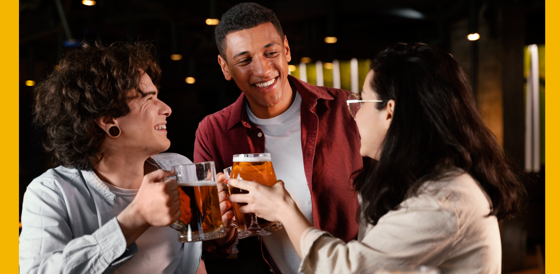 Three friends enjoying drinks together at a bar, smiling and holding beer mugs