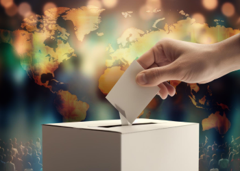 Hand placing a ballot into a box with a world map in the background, symbolizing global elections and democracy.