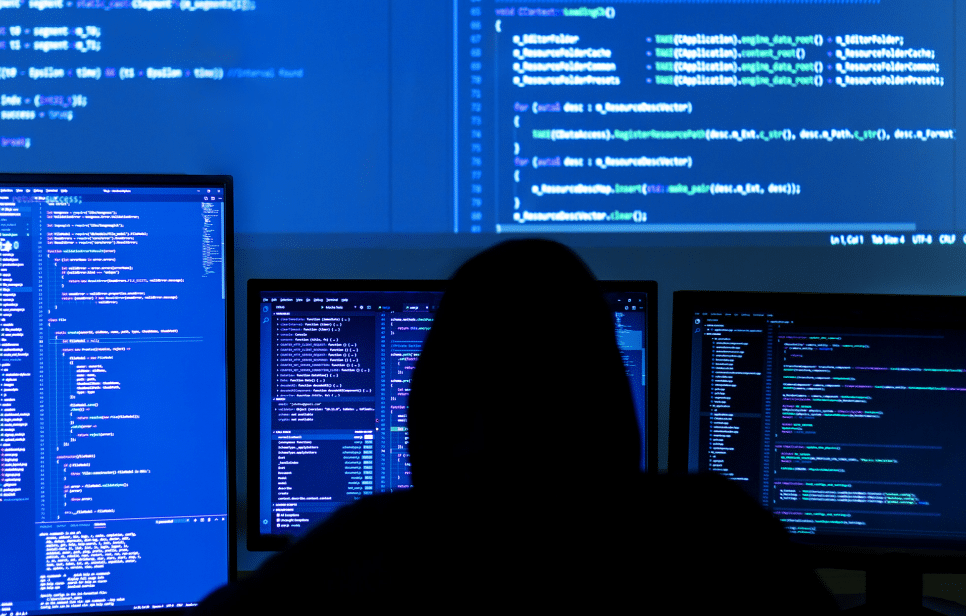 Person in a dark room with multiple computer screens displaying code, representing cybersecurity efforts.