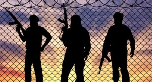 Silhouettes of three ISIS standing in front of a chain-link fence at sunset.